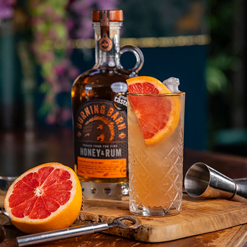 Cocktail in glass with pink grapefruit slice on top, and Burning Barn Honey and Rum bottle in background 