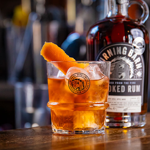 Burning Barn Smoked Rum bottle next to whiskey tumbler filled with cocktail and grapefruit peel on top  